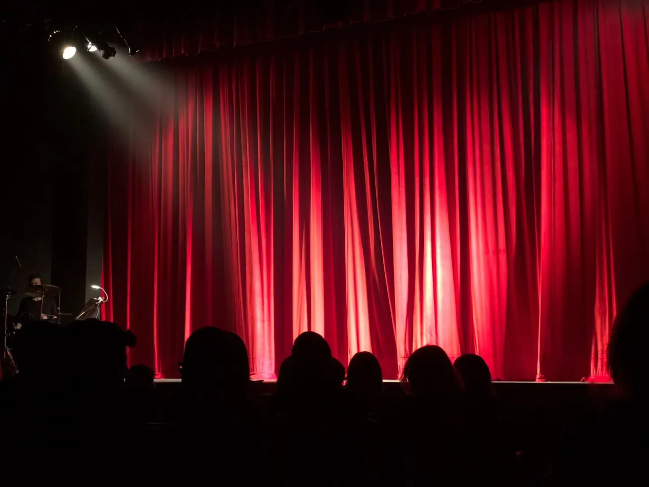 Un teatru cu cortine roșii, reflectoare și siluete ale publicului.
